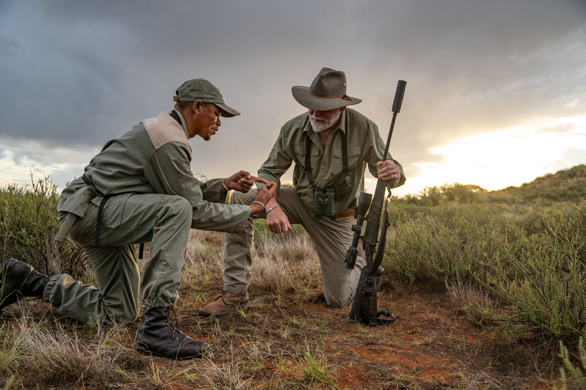 An African tracker advises a hunter who is using a suppressor on his rifle.