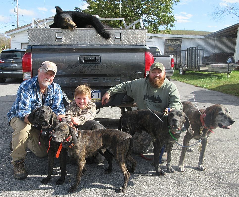 A family of hunters and their dogs kneel in from of a truck with a harvested black bear in the bed.
