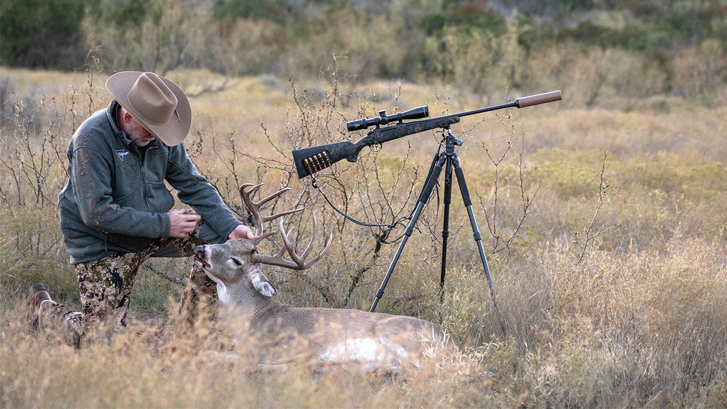 A hunter kneels in a field admiring a harvested whitetail deer with suppressed rifle nearby