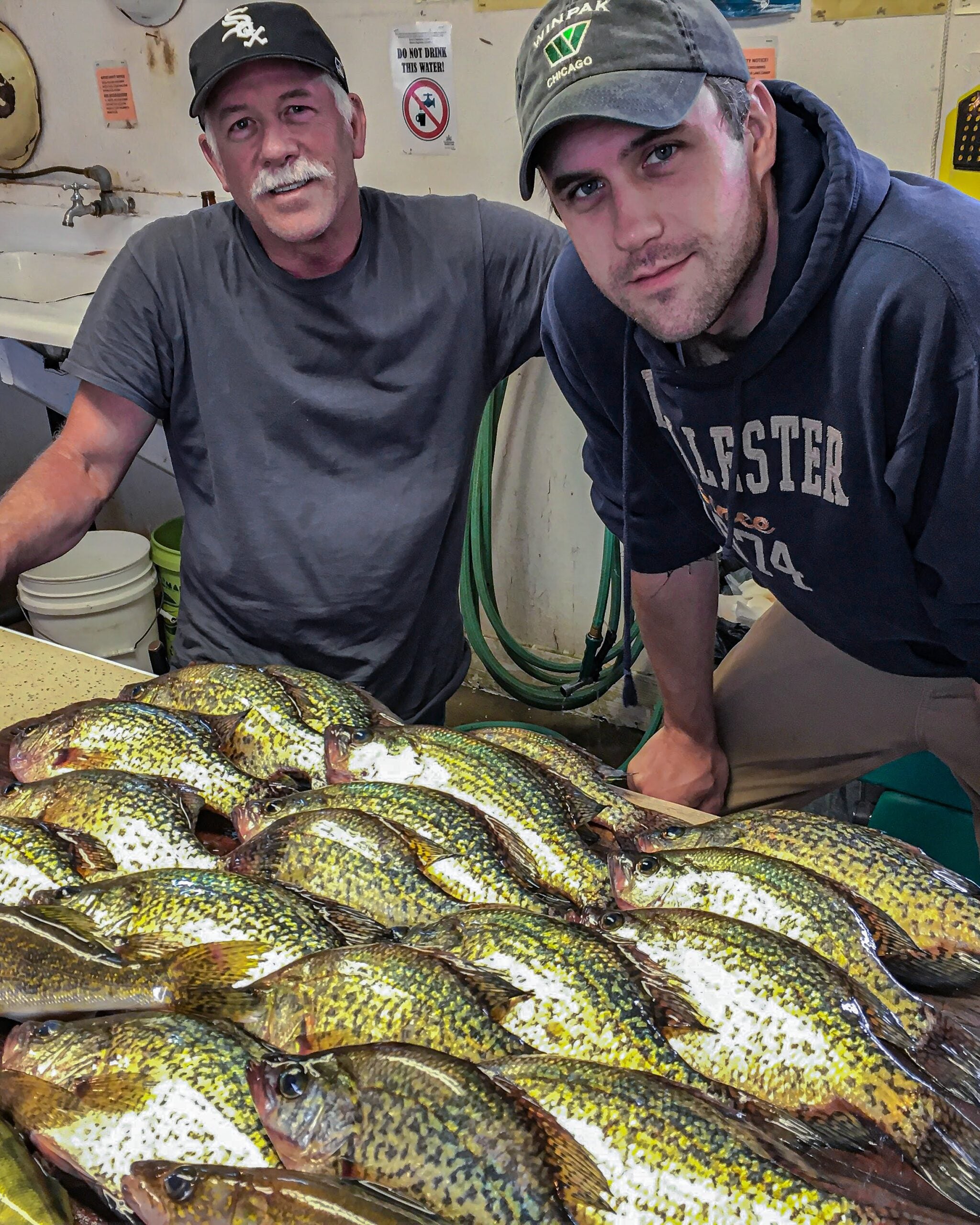 Two fishermen in navy blue shirts stand behind a table of dead crappies.
