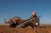 A hunter sits on the ground on the African plain with a wildebeest and rifle.