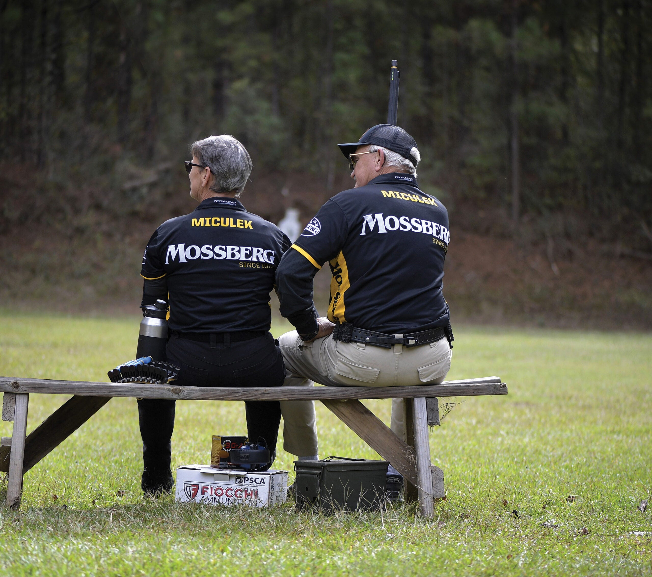 Pro shooters Jerry and Kay Miculek sit on a bench, backs to the camera, at a shooting range.
