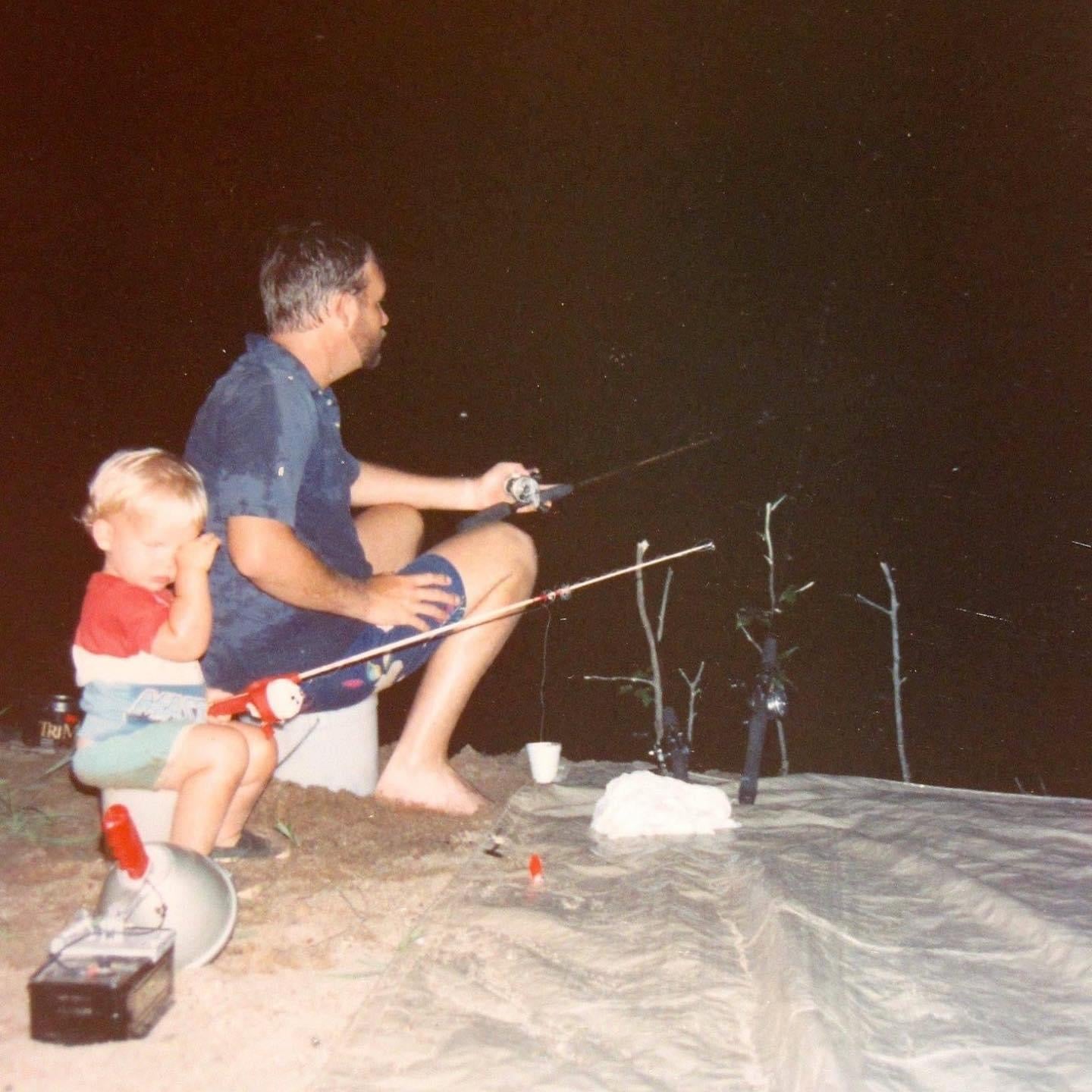 A little boy sits next to his day at night fishing.